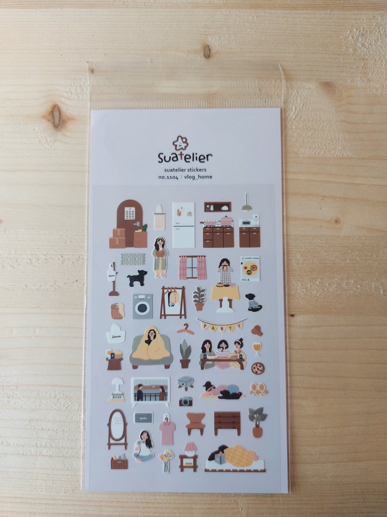 Pu&ograve; includere: Un foglio di adesivi con illustrazioni di un interno di casa, tra cui mobili, piante e persone. Gli adesivi sono su uno sfondo grigio chiaro e hanno una gamma di colori marrone e beige. Il testo "Suatelier" e "no.1104. vlog_home" &egrave; stampato sul foglio.