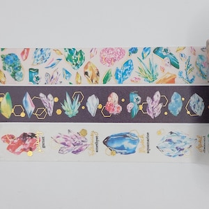 Peut inclure: Trois rouleaux de ruban adhésif washi illustrant des pierres précieuses de différentes couleurs en aquarelle. Le ruban est étiqueté avec les noms des pierres précieuses : grenat, améthyste, aigue-marine et diamant.