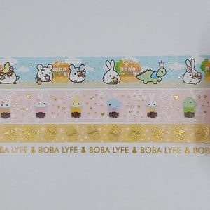 May include: Three rolls of washi tape featuring a pastel color scheme with cute cartoon animals and boba tea. The top roll has a light blue background with white rabbits, a white mouse, and a white bear. The middle roll has a pink background with white rabbits, a yellow chick, a blue whale, and a green cactus. The bottom roll has a yellow background with gold boba tea cups and the text "BOBA LYFE".
