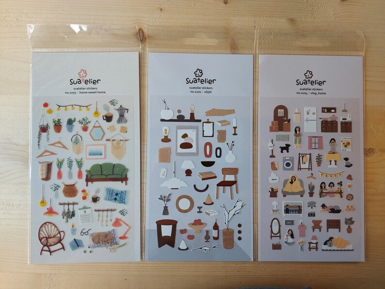 Pu&ograve; includere: Tre fogli di adesivi con illustrazioni di arredamento per la casa, mobili e altri oggetti. Gli adesivi sono in un sacchetto di plastica trasparente e sono etichettati "Suatelier" con il testo "Suatelier stickers no story, home sweet home".