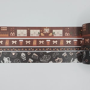 Puede incluir: Un conjunto de cuatro cintas washi marrones y negras con detalles dorados. Las cintas presentan un tema de cafetería con ilustraciones de tazas de café, pasteles y teteras. La cinta superior muestra el interior de una cafetería con mesas y sillas. La segunda cinta tiene un patrón repetitivo de tazas de café con lazos. La tercera cinta tiene un patrón repetitivo de tazas de café, pasteles y teteras. La cinta inferior tiene un patrón repetitivo de tazas de café, pasteles y teteras.