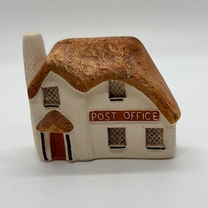 以下が含まれることがあります： 茶色のわらぶき屋根と白い壁を持つ小さな陶器製の小屋。小屋には茶色のドアと黒い枠の窓が2つあります。ドアの上の看板には「POST OFFICE」と書かれています。