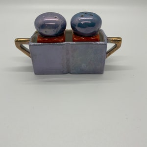 Pode incluir: Um conjunto de saleiro e pimenteiro em um suporte de cerâmica. Os shakers são azuis com um acabamento perolado e têm uma forma redonda. O suporte é lavanda com detalhes dourados e tem uma forma retangular.