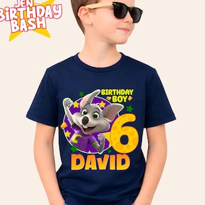 Puede incluir: Camiseta azul marino con un personaje de ratón de dibujos animados y el texto "BIRTHDAY BOY 6 DAVID". La camiseta tiene un número 6 amarillo y estrellas. El texto "JEN BIRTHDAY BASH" tiene forma de estrella.