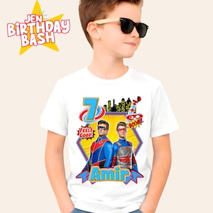 Op de afbeelding: Wit T-shirt met een kleurrijke afbeelding van twee superhelden, het nummer 7 en de naam "Amir". Het ontwerp bevat de zinnen "Feels Good!" en "Boom!" in tekstballonnen. Het shirt maakt deel uit van een verjaardagsthema.