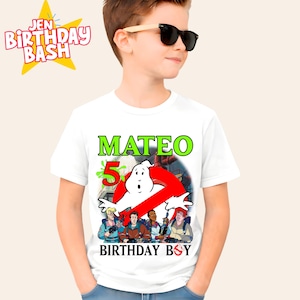 May include: White t-shirt featuring a Ghostbusters-themed design with the name "Mateo" and the number "5" in green. The Ghostbusters logo and characters are also displayed. The text "BIRTHDAY BOY" is at the bottom. The shirt is worn by a person wearing sunglasses.