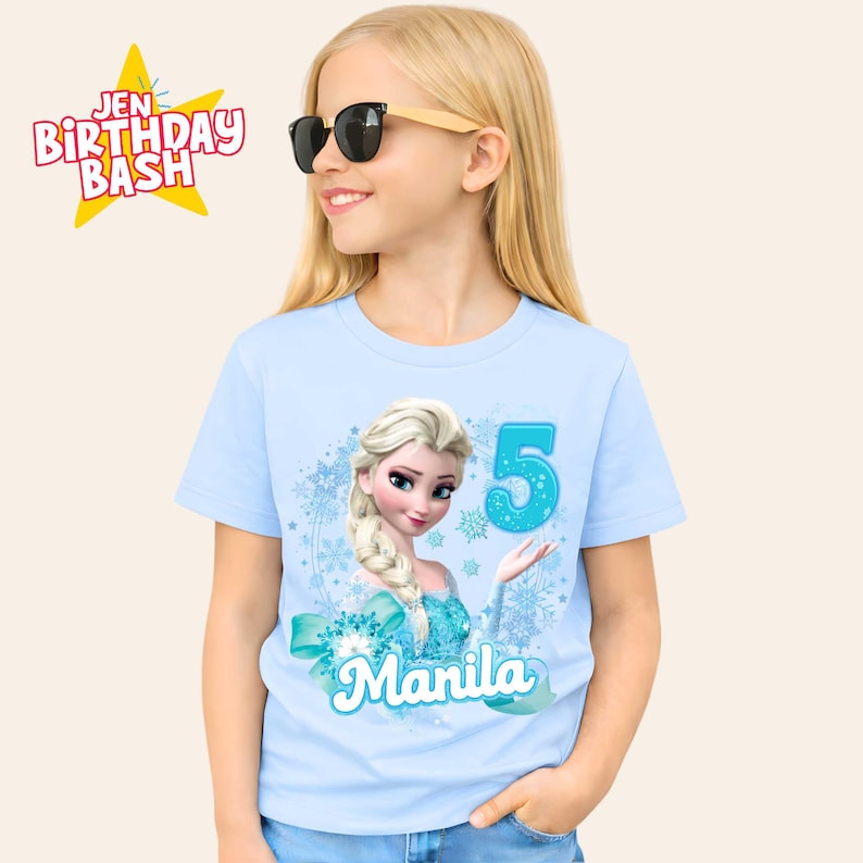 May include: A light blue t-shirt featuring a graphic of Elsa from Frozen, the number 5, and the name "Manila". The shirt also has a teal bow and snowflake accents. The words "Jen Birthday Bash" are in the upper left corner.