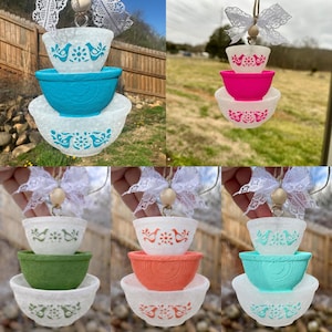 May include: A hanging ornament featuring three stacked bowls of varying sizes, topped with a white lace bow and a wooden bead. The bowls display different color combinations, including teal, pink, green, and orange patterns on white or solid bases.