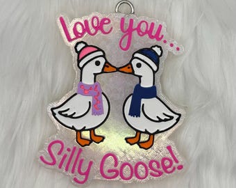 May include: A clear acrylic ornament featuring two cartoon geese kissing. The geese wear winter hats and scarves, with the text "Love you... Silly Goose!" in pink. The ornament has a silver loop for hanging.
