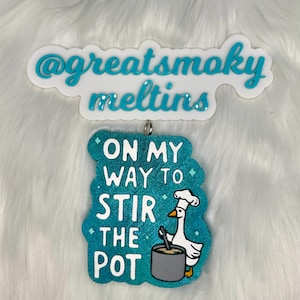 May include: A teal blue ornament with a white goose wearing a chef's hat and stirring a pot. The ornament has the text "On my way to stir the pot" on it.