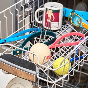 K&ouml;nnte beinhalten: Eine Sp&uuml;lmaschine mit verschiedenen Gegenst&auml;nden darin, darunter eine Tasse, ein Hundehalsband, ein Ball und ein Gummispielzeug. Die Gegenst&auml;nde sind in verschiedenen Farben, darunter blau, rosa, gelb und gr&uuml;n.
