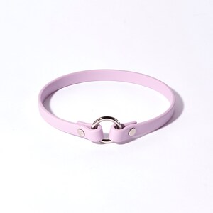 May include: A light purple choker necklace with a silver-colored metal ring and fasteners. The choker is made of a smooth material and is designed to be worn around the neck. The overall design is simple and elegant.