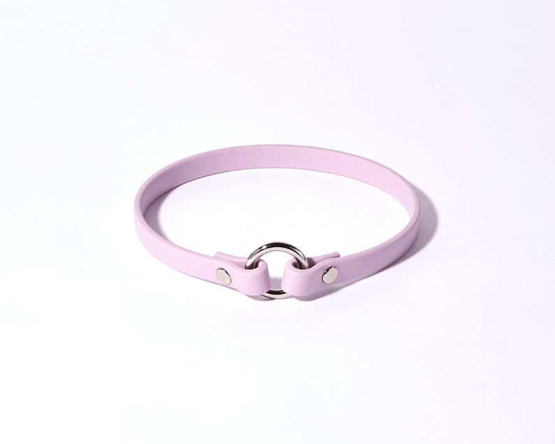 May include: A light purple choker necklace with a silver-colored metal ring and fasteners. The choker is made of a smooth material and is designed to be worn around the neck. The overall design is simple and elegant.