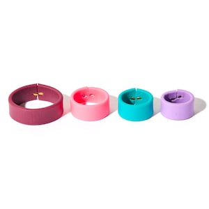 May include: Four colorful silicone ring-shaped objects in varying sizes. The rings are in shades of burgundy, pink, teal, and lavender. Each ring has a small gold-colored accent. The objects are arranged in a row against a white background.