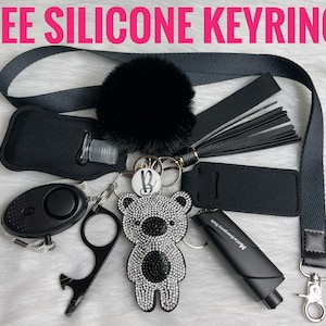 May include: A black and white rhinestone bear keychain with a black tassel, a black hand sanitizer holder, a black personal alarm, a black bottle opener, and a black lanyard with a black pom-pom.
