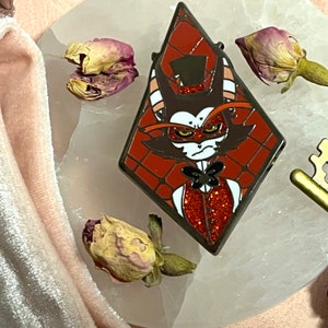 May include: A diamond-shaped enamel pin featuring a cartoon character with a top hat, bow tie, and a red glitter vest. The pin has a red and brown color scheme with black outlines. Dried rose buds and a gold key are in the background.