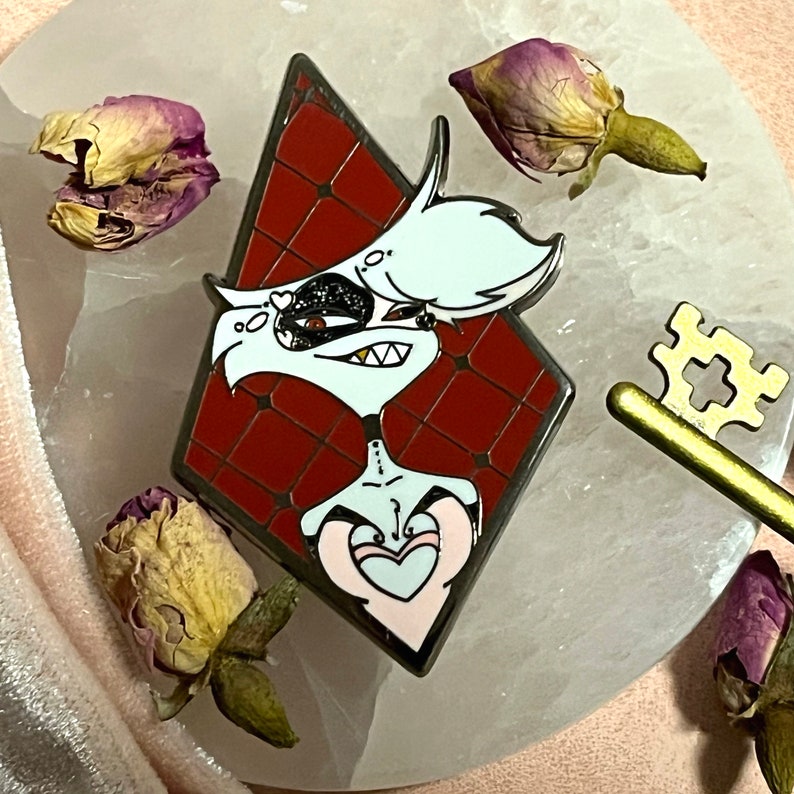 May include: A pin featuring a cartoon character with white hair, a black eye, and a red diamond-shaped background. The pin has a gold outline and is surrounded by dried rose buds and a gold key.