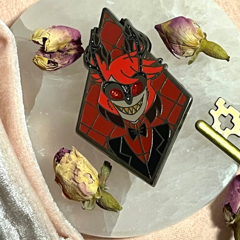 May include: A diamond-shaped enamel pin featuring a cartoon character with red hair, a black suit, and a bow tie. The pin has a red and black color scheme with a black border. Dried rose buds and a gold key are also in the image.