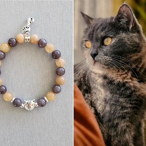 May include: A brown and gray beaded bracelet with a silver cat charm and a silver key charm. The bracelet is on a gray background.
