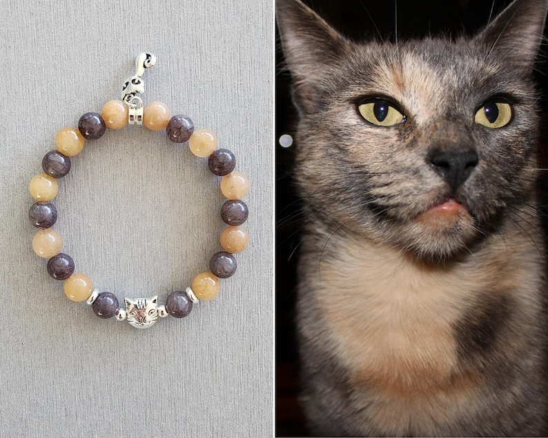 May include: A brown and gray beaded bracelet with a silver cat charm. The beads are round and have a matte finish. The charm is a small, detailed cat head.