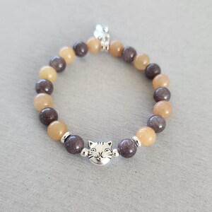 May include: A brown and beige beaded bracelet with a silver cat charm. The bracelet is made of natural stones and is perfect for everyday wear.