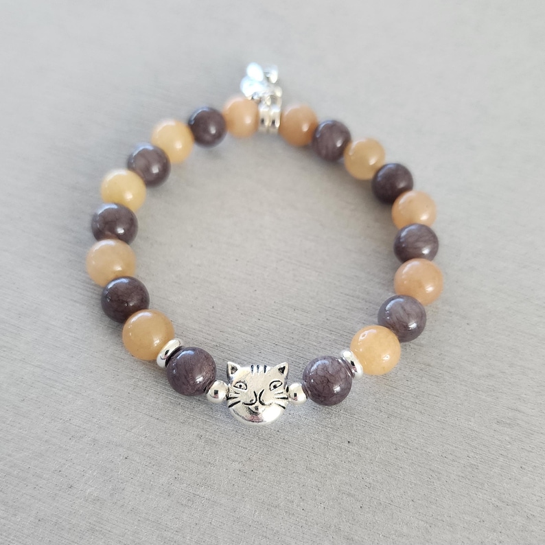 May include: A brown and beige beaded bracelet with a silver cat charm. The bracelet is made of natural stones and is perfect for everyday wear.