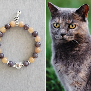 May include: A gray and beige beaded bracelet with a silver cat charm. The bracelet has a silver key charm at the top.