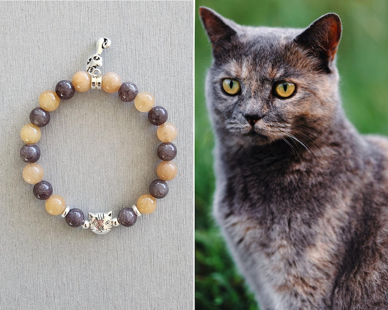 May include: A gray and beige beaded bracelet with a silver cat charm. The bracelet has a silver key charm at the top.