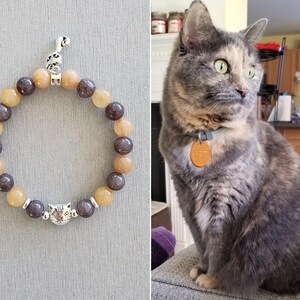 May include: A brown and grey beaded bracelet with a silver cat charm. The bracelet is made of natural stones and has a silver clasp.