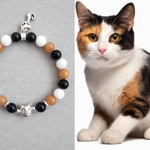 May include: A beaded bracelet featuring alternating black, white, and amber-coloured round beads, accented with a silver-toned metal cat face charm and a small silver-toned cat tail charm, displayed next to a calico cat.