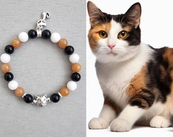 May include: A beaded bracelet featuring alternating black, white, and amber-colored round beads, accented with a silver-toned metal cat face charm and a small silver-toned cat tail charm, displayed next to a calico cat.