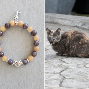May include: A brown and grey cat with green eyes sits on a stone surface. A beaded bracelet with a cat charm is shown in the background.