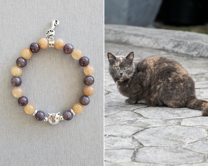 May include: A brown and grey cat with green eyes sits on a stone surface. A beaded bracelet with a cat charm is shown in the background.