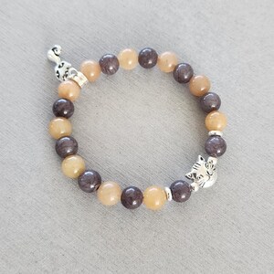May include: A bracelet made of brown and beige beads with a silver cat charm. The bracelet is made of natural stone beads.