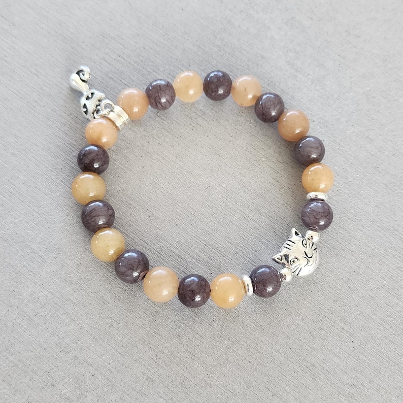 May include: A bracelet made of brown and beige beads with a silver cat charm. The bracelet is made of natural stone beads.