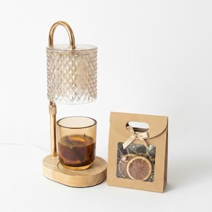 May include: An aromatherapy diffuser with a gold-coloured metal frame and a glass shade. The diffuser sits on a wooden base, with a glass cup containing liquid and spices. A small paper bag with dried orange slices and a bow is next to it.