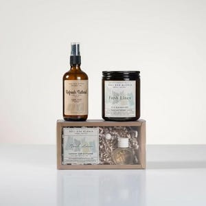 May include: A gift set featuring a brown glass spray bottle, a black jar candle labeled "Fresh Linen", and a small box with a reed diffuser and a small bottle. The set is displayed on a white surface.