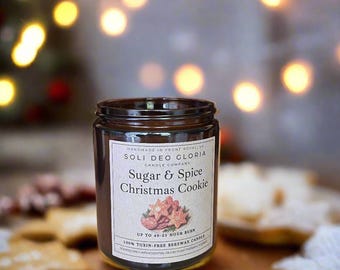 May include: A glass jar candle with a label that reads "Sugar & Spice Christmas Cookie." The candle is surrounded by star-shaped cookies. The label also states "100% Toxin-Free Beeswax Candle" and "Up to 40-55 Hour Burn."