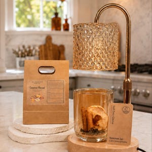 May include: A gold metal candle warmer lamp with a textured glass shade, a wooden base, and a clear glass jar containing dried cinnamon sticks and apple slices. A brown paper bag labelled Cinnamon Harvest and a care instruction card are included.
