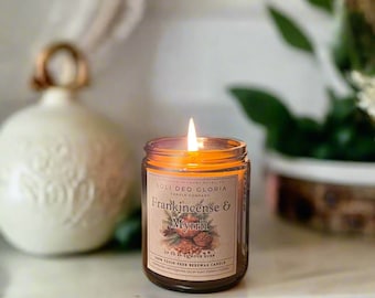 May include: A lit glass jar candle labeled "Frankincense & Myrrh" with a botanical illustration. The candle has a warm glow and is set on a white surface. The label also reads "Soli Deo Gloria Candle Company" and "100% Toxin-Free Beeswax Candle."