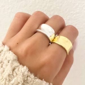 May include: Two rings, one silver and one gold, are stacked on a finger. Both rings are wide and have a curved, rounded shape.