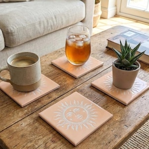 May include: A wooden coffee table displays four square coasters with a sun design, a mug of coffee, a glass of iced tea, and a potted succulent. The coasters are a light peach colour with a white sun graphic.