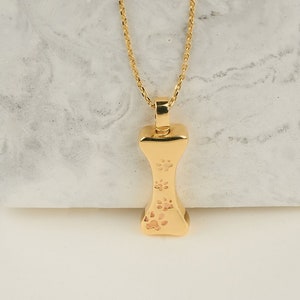 May include: A gold-colored bone-shaped pendant necklace with a delicate chain. The pendant features three paw prints. The necklace is displayed on a white and gray surface, with a black background.