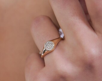 May include: A gold ring with a round, pave-set diamond center. The ring is worn on the finger of a person wearing a white shirt.