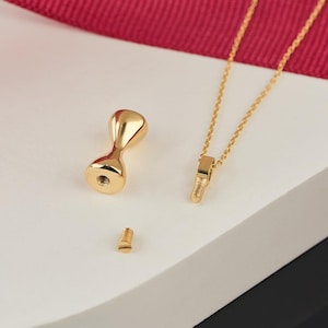 May include: A gold-coloured hourglass-shaped pendant with a screw-on base, a small screw, and a delicate gold chain. The pendant is designed to hold a small amount of cremated remains. The background includes a red ribbon and a white surface.