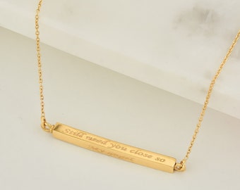 May include: A gold-colored necklace with a rectangular pendant. The pendant has the engraved text "Still need you close so my angel". The necklace chain is delicate and thin. The background is a white marble surface.
