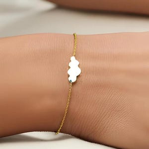 May include: A delicate gold-colored bracelet featuring a small, white, cloud-shaped charm. The bracelet is worn on a wrist, showcasing the minimalist design and fine details of the jewelry. The charm is approximately 0.5 inches in length.