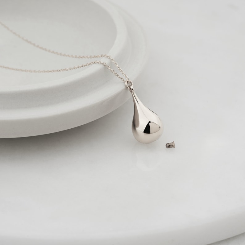 Sterling silver teardrop cremation urn necklace with tiny screw cap shown beside it on a white ceramic display
