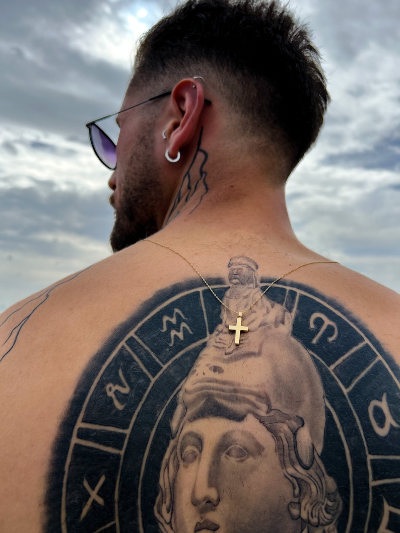 May include: A person with a black and white tattoo of a zodiac wheel on their back. The tattoo features a woman's face, astrological symbols, and a small gold cross pendant.