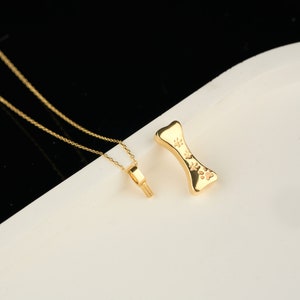 May include: A gold-colored necklace with a small pendant and a separate bone-shaped charm. The bone charm has paw prints. The necklace and charm are displayed on a white surface with a black background.